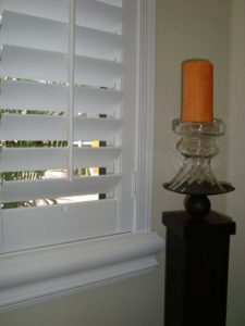 White plantation shutters with a tall wooden candle stand displaying a orange candle