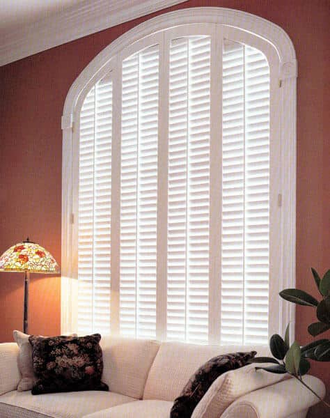 A large arched custom plantation shutter window that has white shutters and a wide trim moulding with hinges on each side
